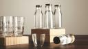 Perfect set of pure clean healthy water in transparent glass bottles and cups presented on wooden plates on tableOne cup and bottle in front close up