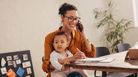 Businesswoman talking on phone with her baby sitting on her lap at home. Working mother making notes while talking on mobile phone.