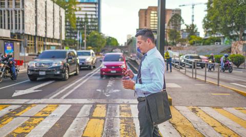 A young man crossing the street of a big city with a cell phone and a  coffee cup in his hands.