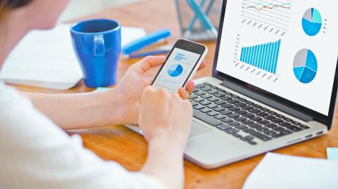 Businesswoman holding smartphone while working with laptop at home office desk, using cross platform responsive design apps for project analysis, analyzing cloud data statistics, close up rear view