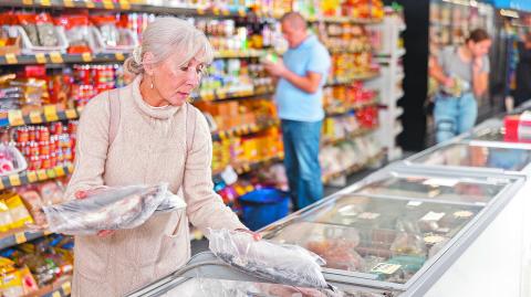 Interested slender elderly woman with silver hair pulled back into ponytail wearing casual knitted dress shopping in grocery store, choosing frozen fish in glass refrigerated display case