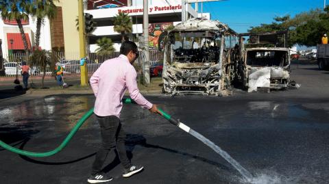 Un hombre limpia una calle junto a vehículos quemados tras diversos ataques del crimen organizado.