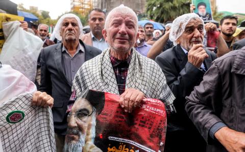 Un hombre llora mientras sostiene un cartel que dice "cruzando una línea roja" en persa junto a una imagen de Hassan Nasrallah.