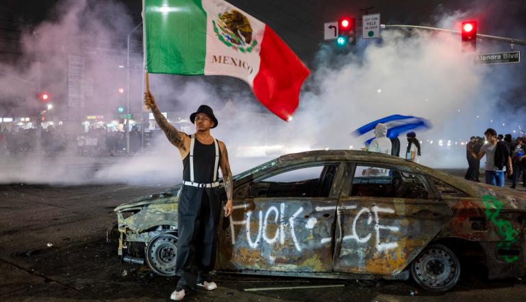 Un manifestante ondea la bandera mexicana junto a los restos de un auto quemado, pintado con grafitis contra el Servicio de Inmigración y Control de Aduanas de Estados Unidos (ICE).