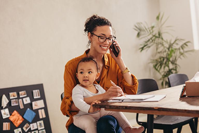 Businesswoman talking on phone with her baby sitting on her lap at home. Working mother making notes while talking on mobile phone.