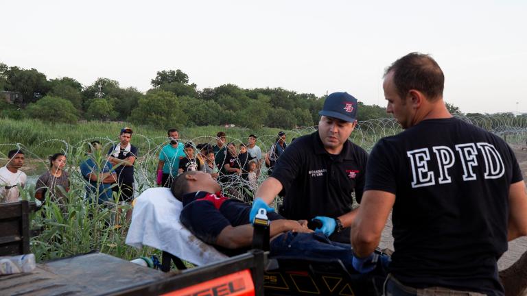 Migrants watch from behind rows of razor wire as firefighter EMTs Rudy Castillon and William Dorsey transfer a Venezuelan man experiencing fatigue, abdominal pain and vomiting while crossing the Rio Grande River, onto a stretcher, in Eagle Pass, Texas, U.S., July 11, 2023. REUTERS/Kaylee Greenlee Beal