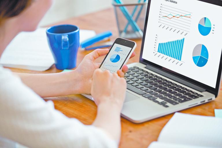 Businesswoman holding smartphone while working with laptop at home office desk, using cross platform responsive design apps for project analysis, analyzing cloud data statistics, close up rear view