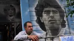 A man stands next to a mural depicting late Argentine football legend Diego Maradona outside the Diego Armando Maradona Stadium, home of Argentine football team Argentinos Juniors at La Paternal neighborhood in Buenos Aires on December 2, 2025. A court in Buenos Aires will decide on Tuesday, during a preliminary hearing how a new trial will be conducted for the death of Diego Maradona in 2020, following the annulment of the first trial. The scheduled start date has been set for March 17, 2026, although appeals by the defence teams of the seven accused health professionals still need to be addressed, which could delay the start. (Photo by Luis ROBAYO / AFP)
