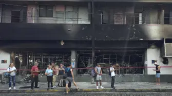 People stand next to a burned building in Puerto Vallarta, Jalisco state, Mexico, on February 23, 2026. Mexico has deployed 10,000 troops to quell clashes sparked by the killing of the country's most wanted drug lord, which have left dozens dead, officials said on February 23. Nemesio "El Mencho" Oseguera, leader of the Jalisco New Generation Cartel (CJNG), was wounded on February 22 in a shootout with soldiers in the town of Tapalpa in Jalisco state and died while being flown to Mexico City, the army said. (Photo by Arturo MONTERO / AFP)