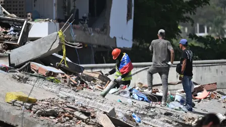 A man wrapped in a Venezuelan national flag tries to find missing people amid the rubble at Los Cocos beach, in Caraballeda, La Guaira state, Venezuela, on July 1, 2026, following the June 24 twin earthquakes. Venezuelans, with help from around the world, were still searching on July 1, 2026 for survivors a week after massive earthquakes killed nearly 2,000 and left thousands unaccounted for. (Photo by Juan BARRETO / AFP)