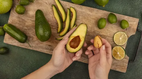 top view of female hands holding an avocado in one hand and its pit in the other hand on a wooden kitchen board with limes feijoas and green apples isolated on a green background