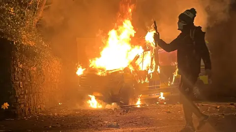 Un manifestante filma y un vehículo policial arde de fondo.