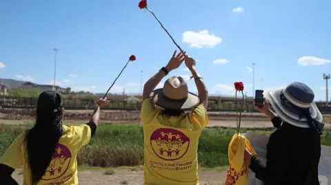 Personas saludan con flores rojas durante la protesta binacional "Madres de la Frontera: Amor sin fronteras".