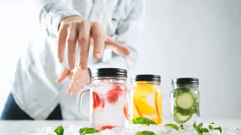 Hand takes one of rustic jars with cold fresh homemade lemonades from strawberry, orange, cucumber, ice and mint Close focus on first jar, action move, summer mood