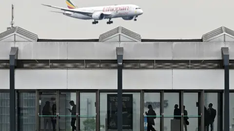People wait for a train at a metro station as an Ethiopian Airlines Boeing 787-8 passenger plane flies in the sky during landing at the Copenhagen Airport in Copenhagen, Denmark, on June 4, 2025. (Photo by Sergei GAPON / AFP)