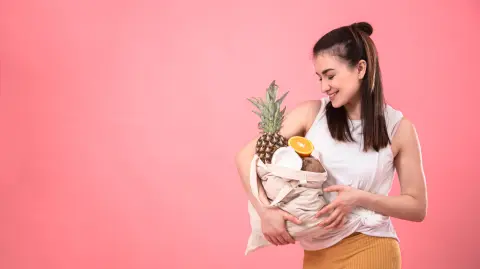 Stylish young girl smiling and holding an eco bag with exotic fruits on a pink background copy space.