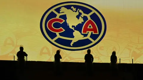 Silhouettes of riot police officers in front of the jumbotron showing the America football club badge ahead of the Liga MX Clausura football match between America and Cruz Azul at the Sports City Stadium in Mexico City on April 12, 2025. (Photo by Alfredo ESTRELLA / AFP)