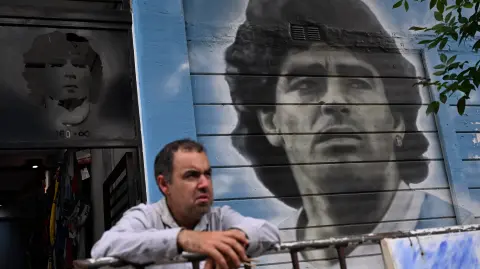 A man stands next to a mural depicting late Argentine football legend Diego Maradona outside the Diego Armando Maradona Stadium, home of Argentine football team Argentinos Juniors at La Paternal neighborhood in Buenos Aires on December 2, 2025. A court in Buenos Aires will decide on Tuesday, during a preliminary hearing how a new trial will be conducted for the death of Diego Maradona in 2020, following the annulment of the first trial. The scheduled start date has been set for March 17, 2026, although appeals by the defence teams of the seven accused health professionals still need to be addressed, which could delay the start. (Photo by Luis ROBAYO / AFP)