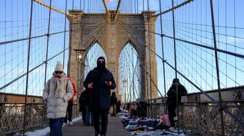 People brave the cold temperatures while walking on the Brooklyn Bridge in the Manhattan borough of New York City on January 21, 2026. Bringing icy temperatures, Winter Storm Fern will slam a massive stretch of the United States this week, with more than 175 million people facing the prospect of heavy snowfall, power outages and travel disruption. Forecasters warned it could be 2,000 miles (3,219 kilometers) long, well over half the length of the continental US. (Photo by CHARLY TRIBALLEAU / AFP)