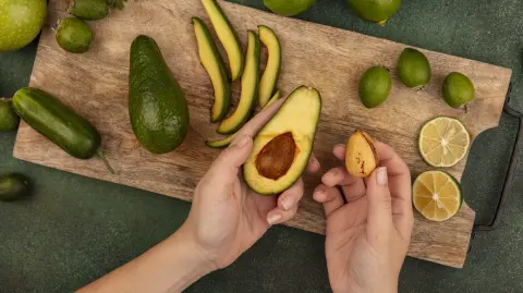 top view of female hands holding an avocado in one hand and its pit in the other hand on a wooden kitchen board with limes feijoas and green apples isolated on a green background
