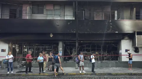 People stand next to a burned building in Puerto Vallarta, Jalisco state, Mexico, on February 23, 2026. Mexico has deployed 10,000 troops to quell clashes sparked by the killing of the country's most wanted drug lord, which have left dozens dead, officials said on February 23. Nemesio "El Mencho" Oseguera, leader of the Jalisco New Generation Cartel (CJNG), was wounded on February 22 in a shootout with soldiers in the town of Tapalpa in Jalisco state and died while being flown to Mexico City, the army said. (Photo by Arturo MONTERO / AFP)