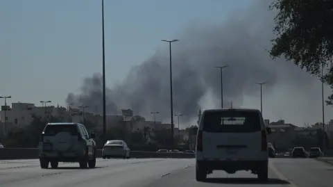 Motorists drive along a street as smoke rises from a reported Iranian strike in the area where the US Embassy is located in Kuwait City on March 2, 2026. Black smoke was seen rising from the US embassy in Kuwait City on March 2 after the latest volley of Iranian strikes, an AFP correspondent saw. (Photo by AFP)