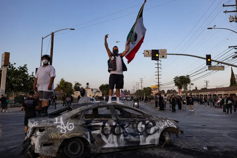 Un manifestante con una bandera mexicana se encuentra de pie sobre un auto quemado durante un enfrentamiento con las fuerzas del orden.